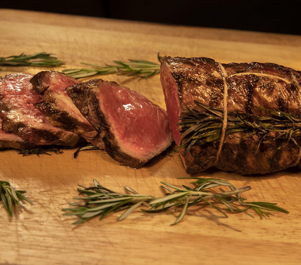 Slices of roast beef on a wood cutting board surrounded by rosemary