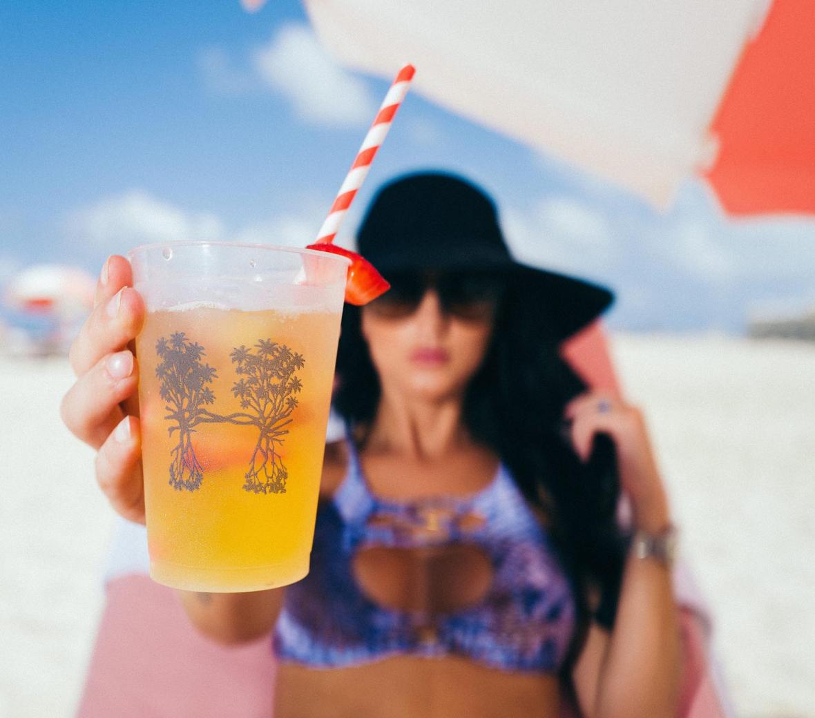 a woman holding a drink up to the camera while sitting on the beach