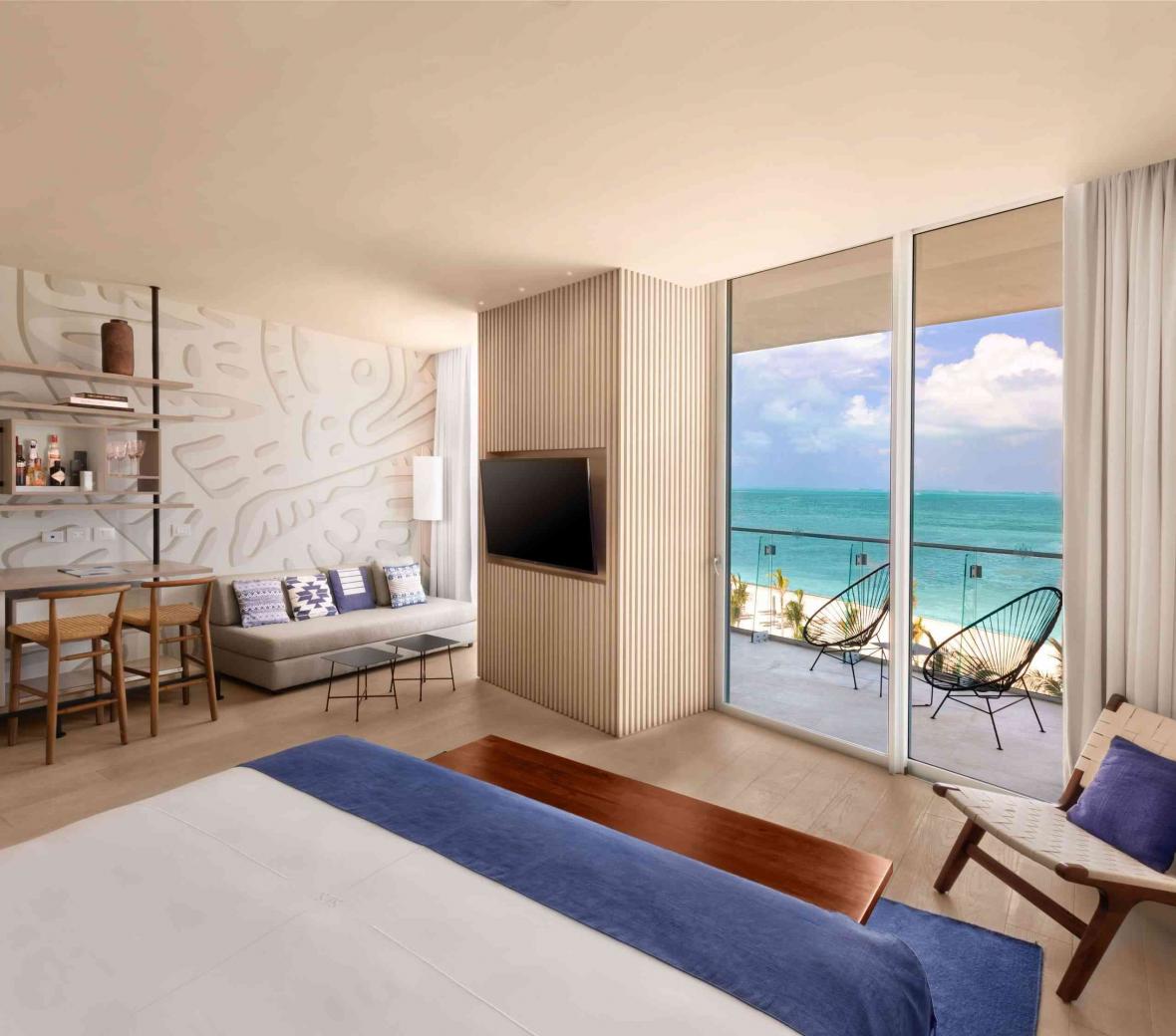 Wide shot of the Deluxe Ocean Front King Room that includes a desk, sitting area, television, and a balcony with a view of the ocean