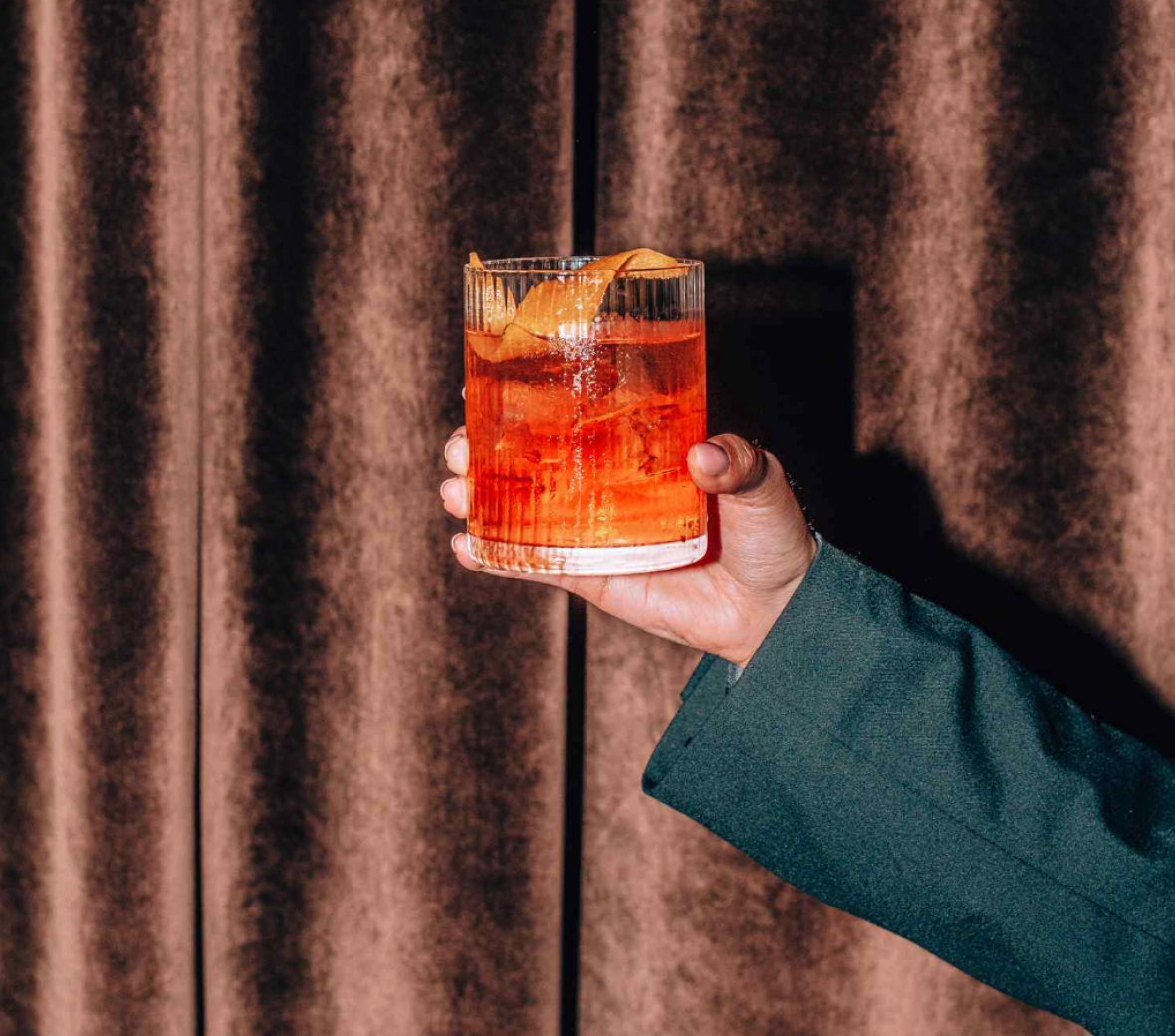 A man holds a festive orange cocktail, it looks like a Negroni