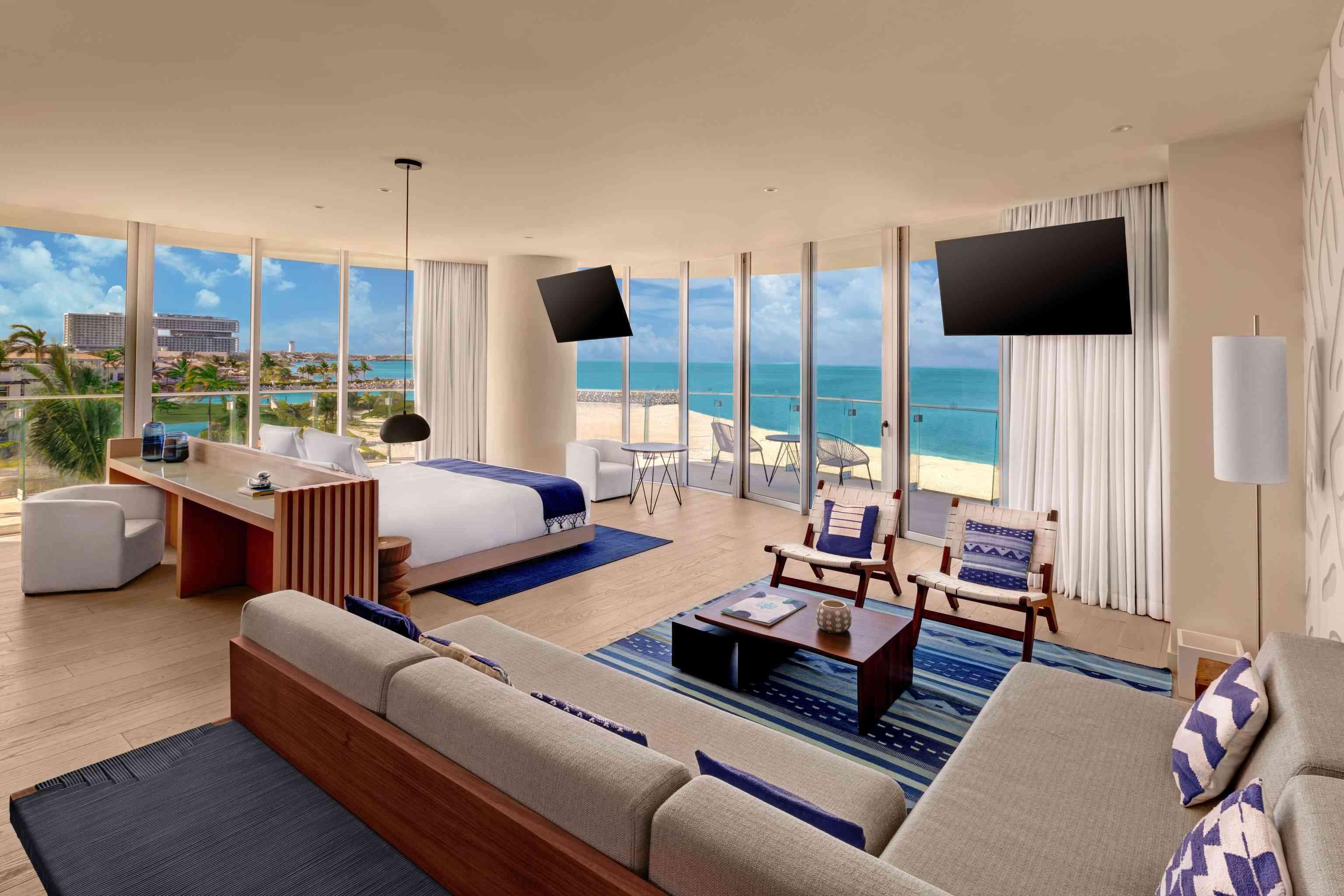 Wide shot of the Premier Ocean Front One Bedroom Suite that includes a desk, a large seating area, 2 televisions, a bed, and a balcony with a view of the ocean