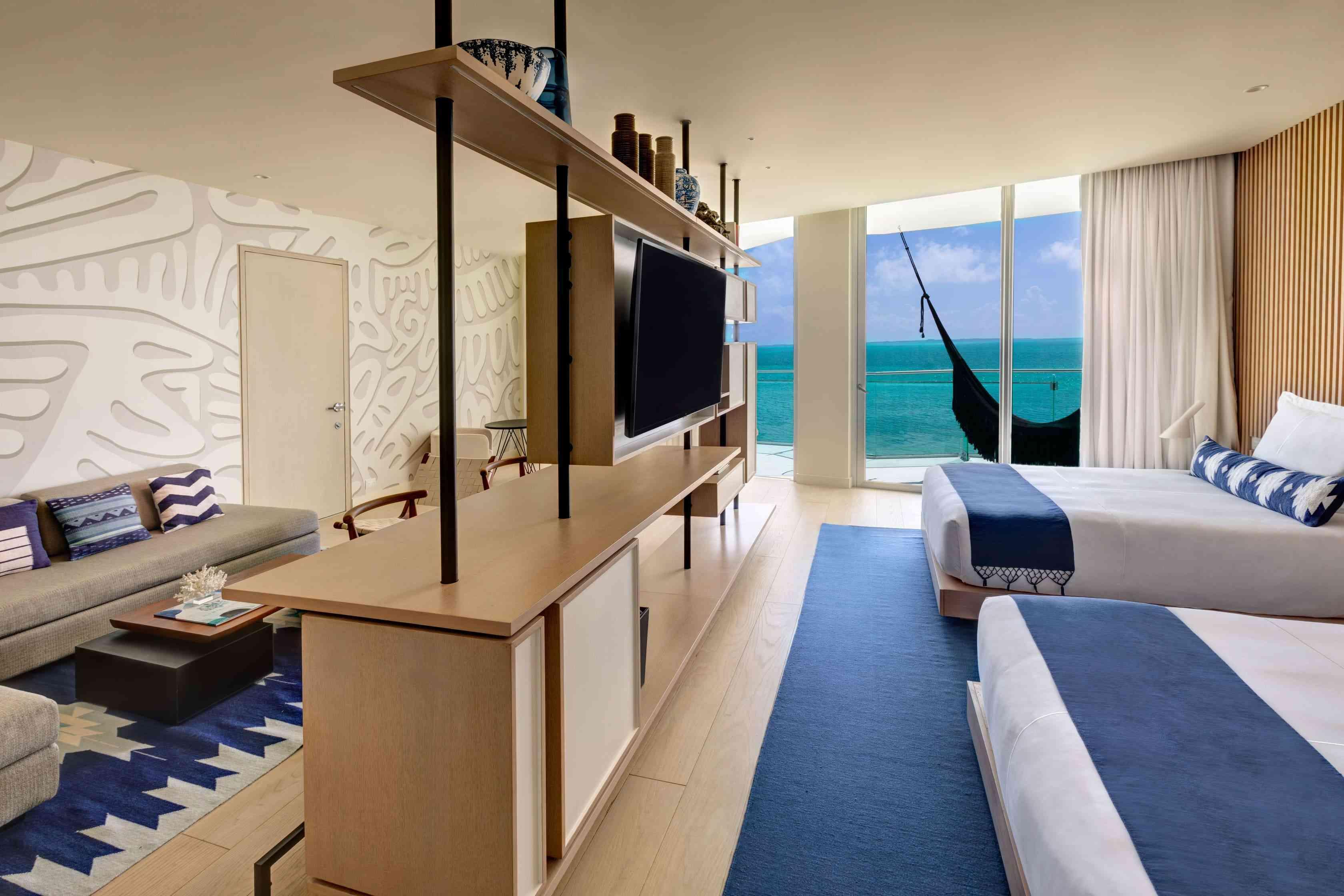 Shot of the Deluxe Ocean Front One Bedroom Suite which includes two double beds, a television, a desks that separates the bedroom from the living room, and a balcony with a hammock overlooking the ocean